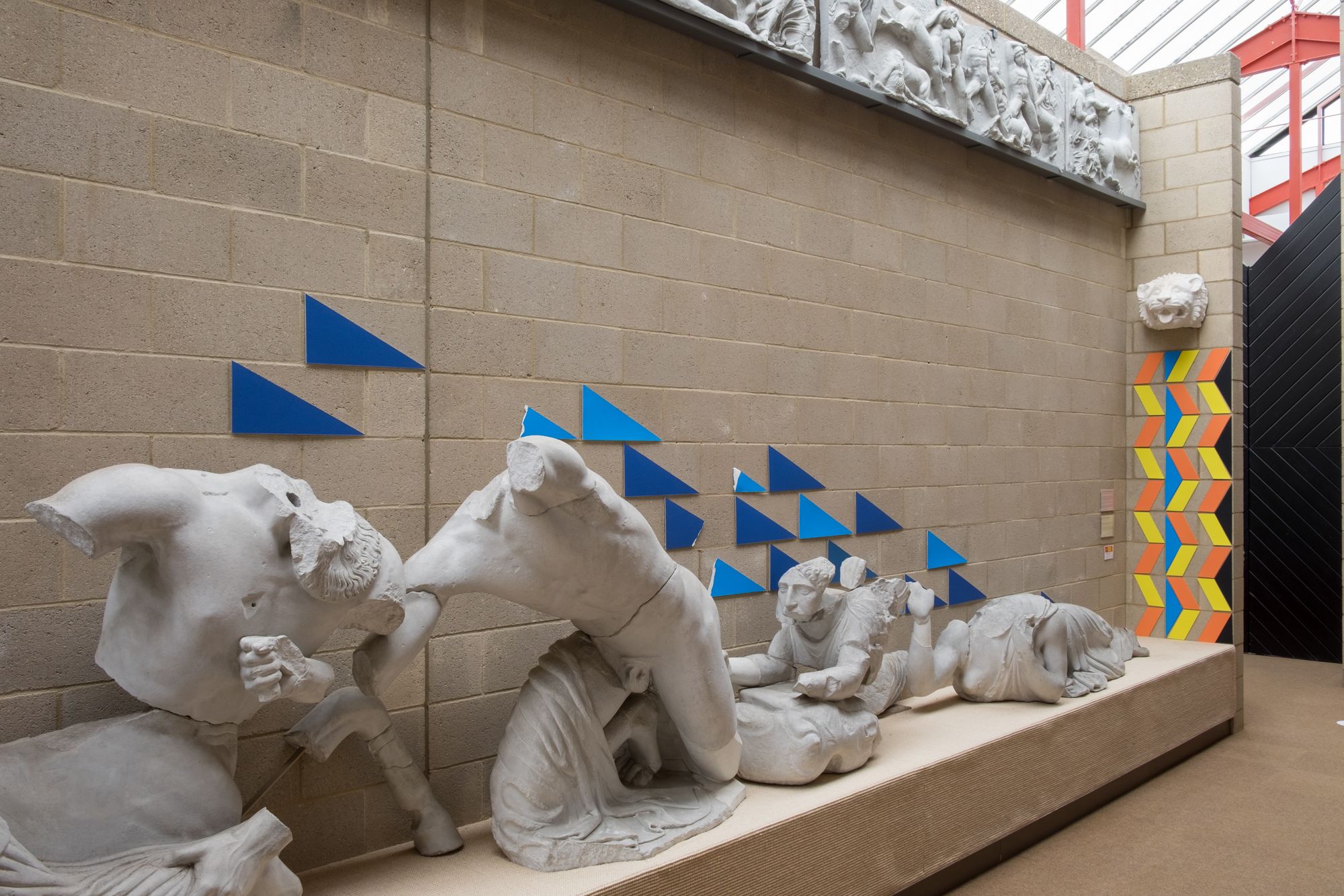 Plaster Casts figures from a pediment. Blue triangles mounted on the wall slope down in line with the figures. To the right colourful geometric patterns go up the wall.