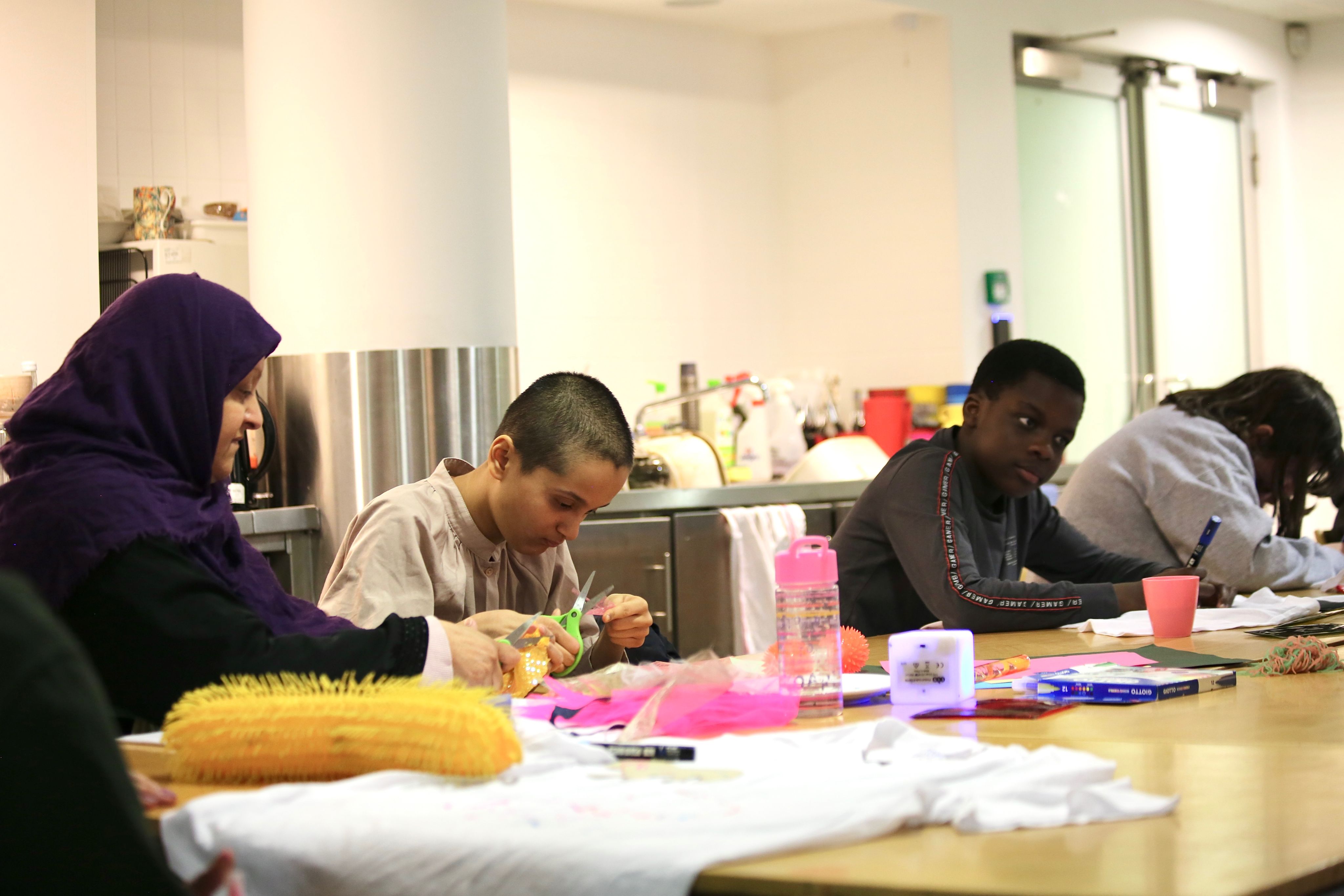 Khadija leans over a wooden table sitting next to her mother on the left and other participants on the right snipping at collage materials with a pair of scissors.