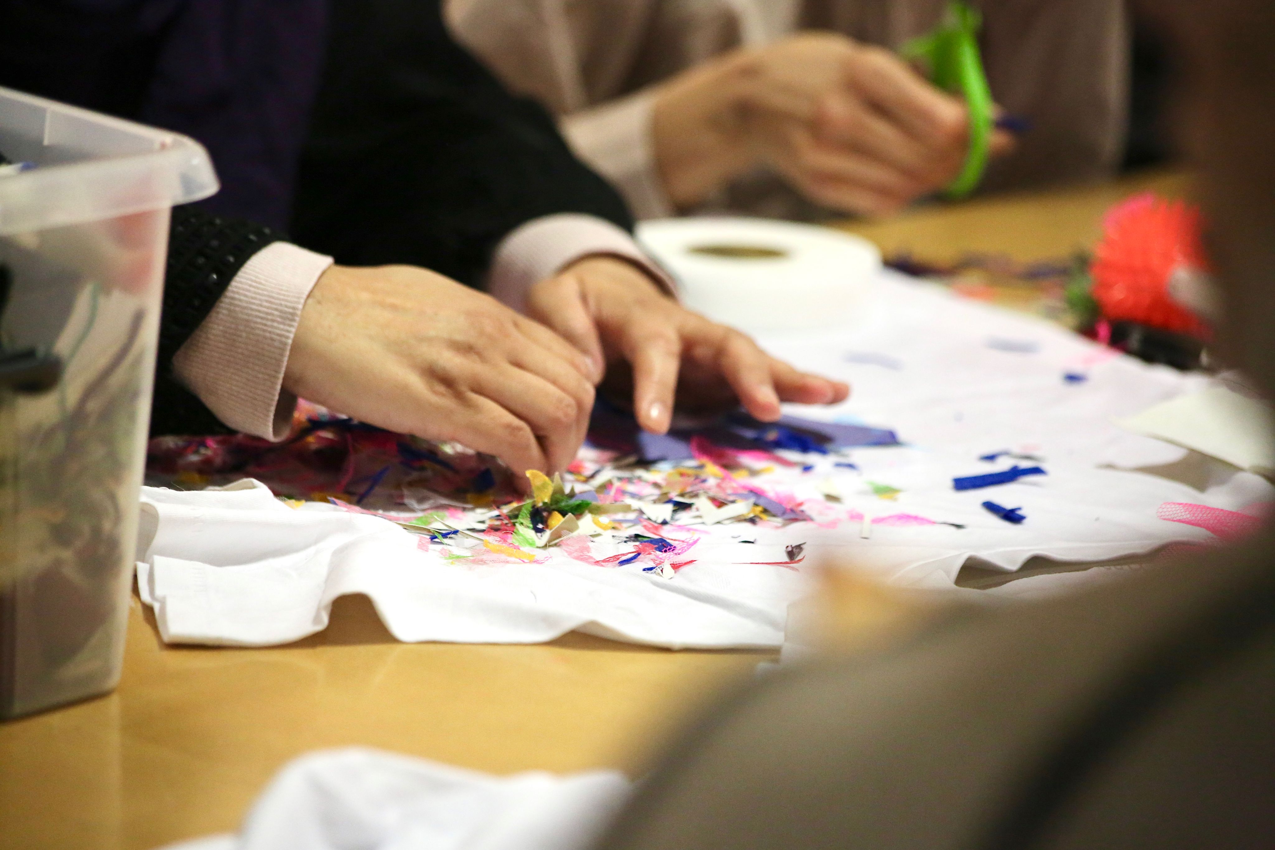 Sima's hands creating a colourful collage on a white t-shirt on a wooden table.