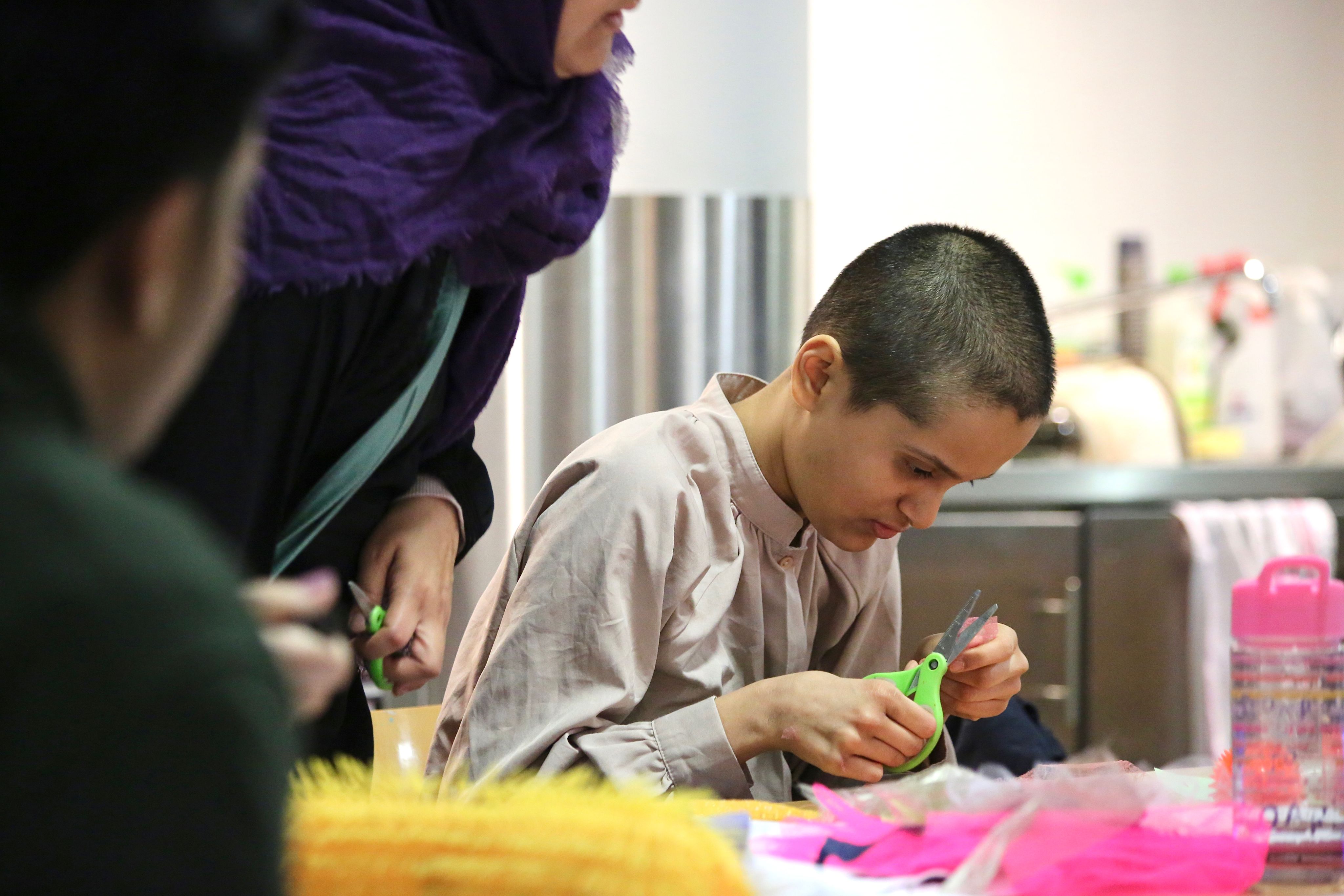 Khadija leans over a wooden table sitting next to her mother on the left and other participants on the right snipping at collage materials with a pair of scissors.