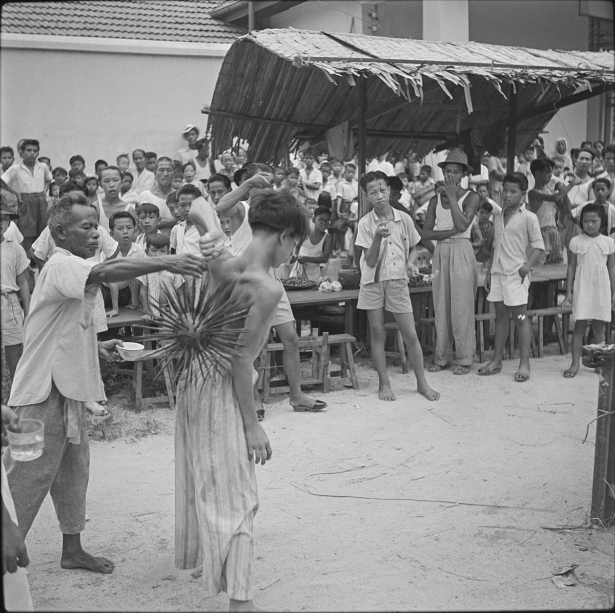 In a street, in front of a wooden stall with thatch roof, an older man wearing a shirt and trousers, supports a young man as he hits his bare back with a ball of spikes. A group of men, women and children watch from the roadside.