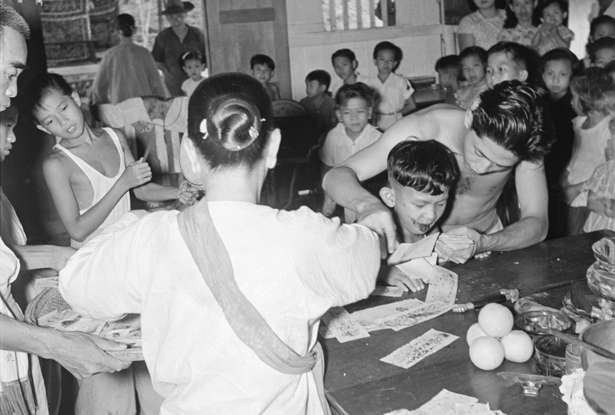 Inside a Chinese temple, the spirit-medium, a boy, leans forward dripping blood from his tongue onto a charm paper held by an assistant. More papers lie on the offering table in front of him. In the foreground is an older woman wearing a white blouse and sash over her left shoulder. A group of onlookers are in the background.