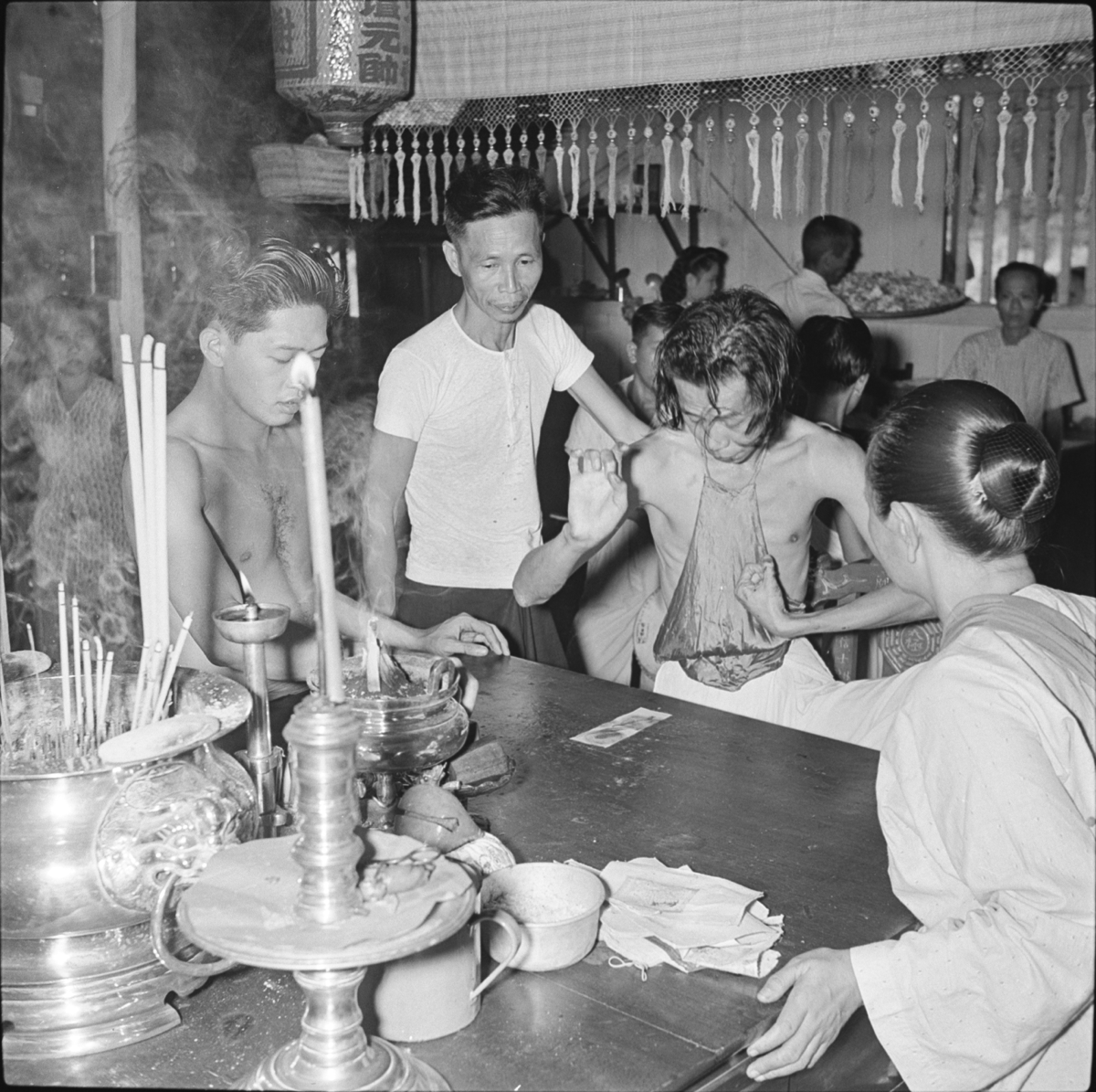 The spirit-medium, a young man with dark hair, wearing a white apron and silk stomacher, gestures with his hands as he looks down at a charm paper on the wooden offering table in front of him. Beside him is an older man in a white t-shirt, a young man and an older woman in a white blouse. On the table are lighted candles and incense sticks, bowls of incense and further charm papers.