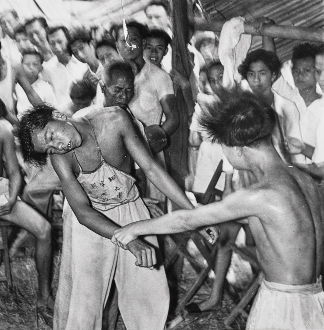 Inside a temple a young man holds the wrists of a second young man, dressed in loose trousers and a stomacher, who appears to be in a trance state. Onlookers watch from outside the temple.