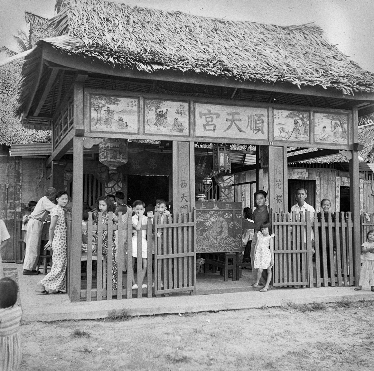 In the porch are a number of people, and a painted sign balancing on benches. The temple has a thatch roof, painted figures and scenes over the porch.