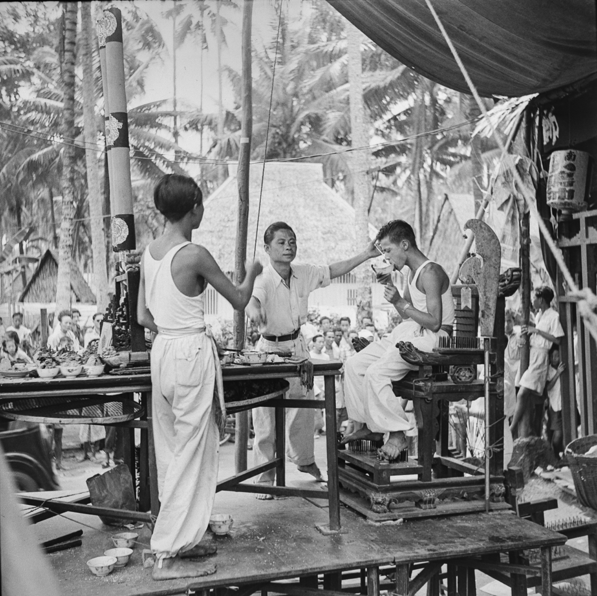 A young man wearing a white vest and trousers sits on a "spike chair" holding a bowl to his lips. Two older men assist, one holding a spike piercing his lip. A large crowd watches.