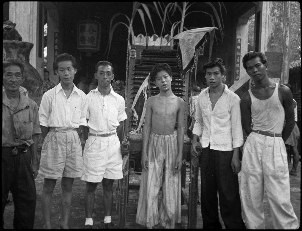 The dang-ki, a young man wears light-coloured trousers. Two of his assistants wear white shirts and shorts, a third wears white shirt and dark trousers, the fourth a white vest and trousers.
