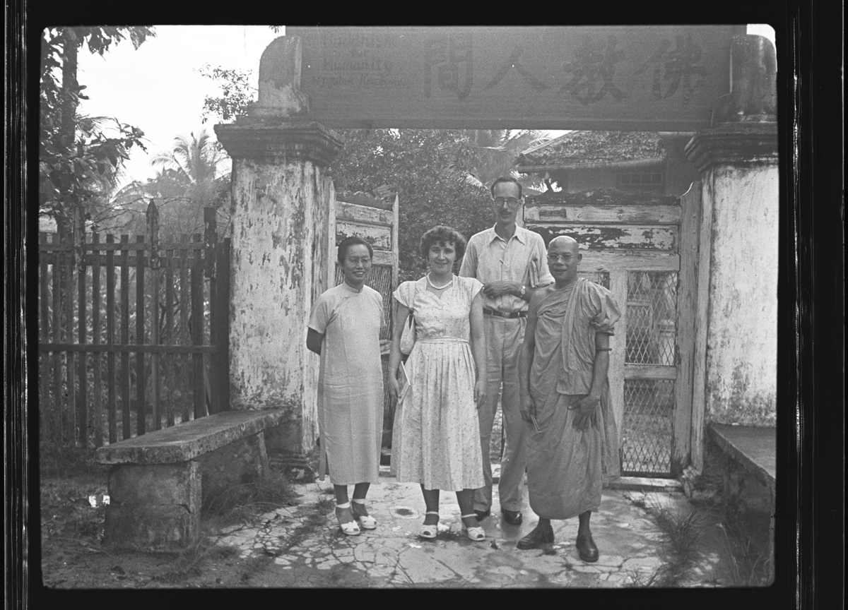 A man is standing in front of an entrance gateway with a European woman, a Chinese woman, and a Chinese monk. A sign in Chinese, above the open double gate reads: 'Buddhism for humanity'.
