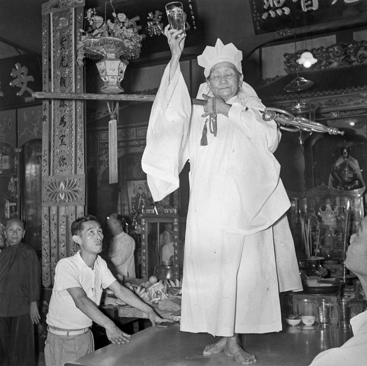 The Dang-ki, an older woman, wears a white five-pointed headdress and white robe. She is standing on an offering table holding a wooden staff or key horizontally across her body. In her right hand she holds up a glass of water. On the left, a man stands with hands on the table looking up at the Dang-ki.