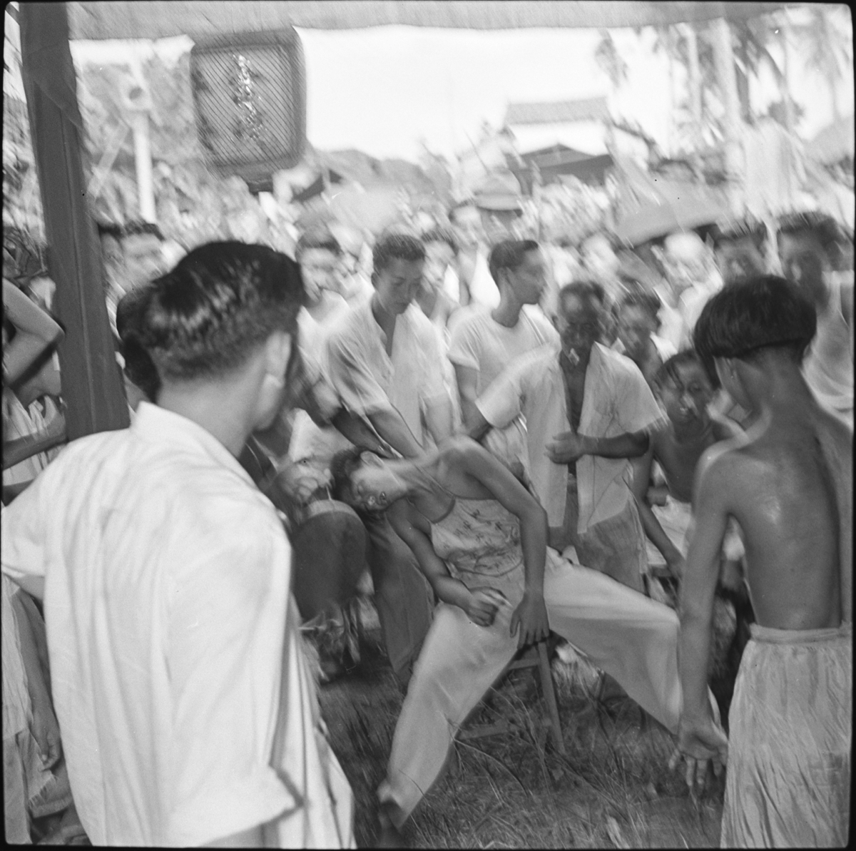 Under the temple awning, a young spirit medium sits on a wooden chair, slumped to one side. A group of onlookers and a second spirit-medium stand watching. A paper lantern hanging from the awning has Chinese characters which may give the name of the temple or deity.