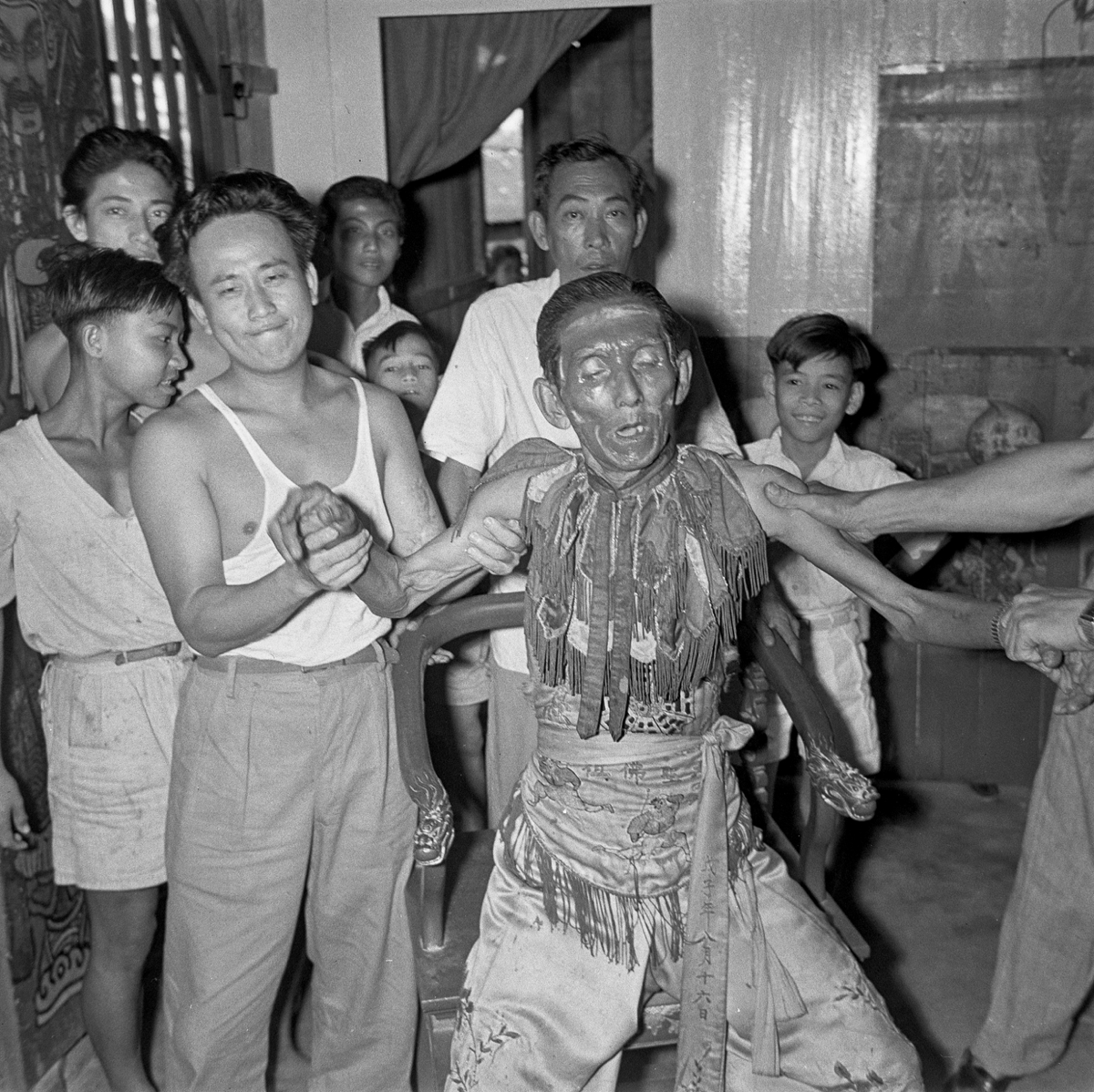 The an older spirit medium has his face painted and wears a collar and silk trousers. He is standing at the front with knees slightly bent, probably in a trance state with his eyes closed. On his left and right, younger men (cropped by the frame on the right) support his arms. A small group of onlookers stand behind the medium.