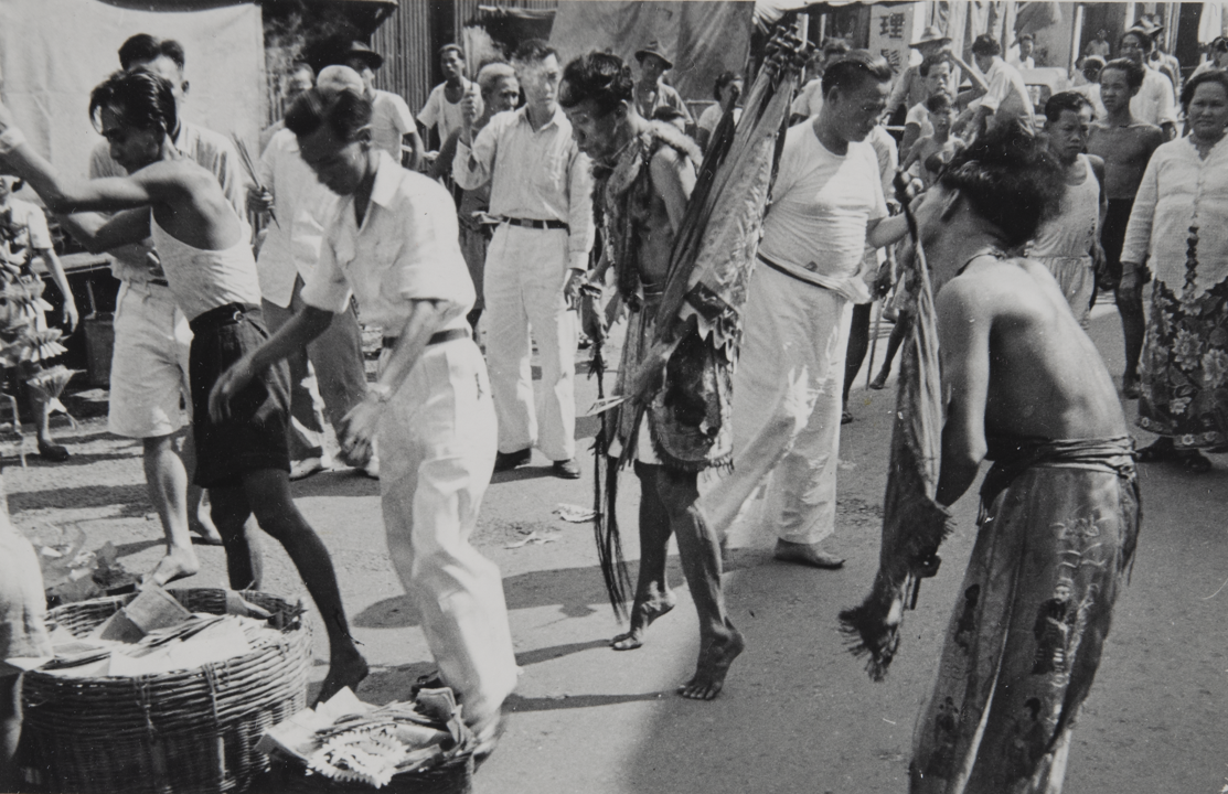 One medium, a middle-aged man, stands on tiptoes in the middle of the street, looking downwards. In his left hand are flags, and in his right hand an object, possibly a sword. On the right a second, younger medium wears trousers and an apron. He holds a flag in his left hand and his head inclined to the right. On the left, two male assistants reach towards baskets filled with printed papers.