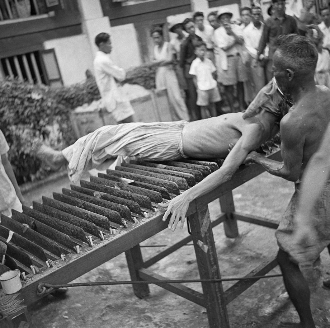An assistant supports the shoulders of a dang-ki spirit medium, an older man wearing cloth headdress and loose trousers, who is lying supine on a bed of blades.