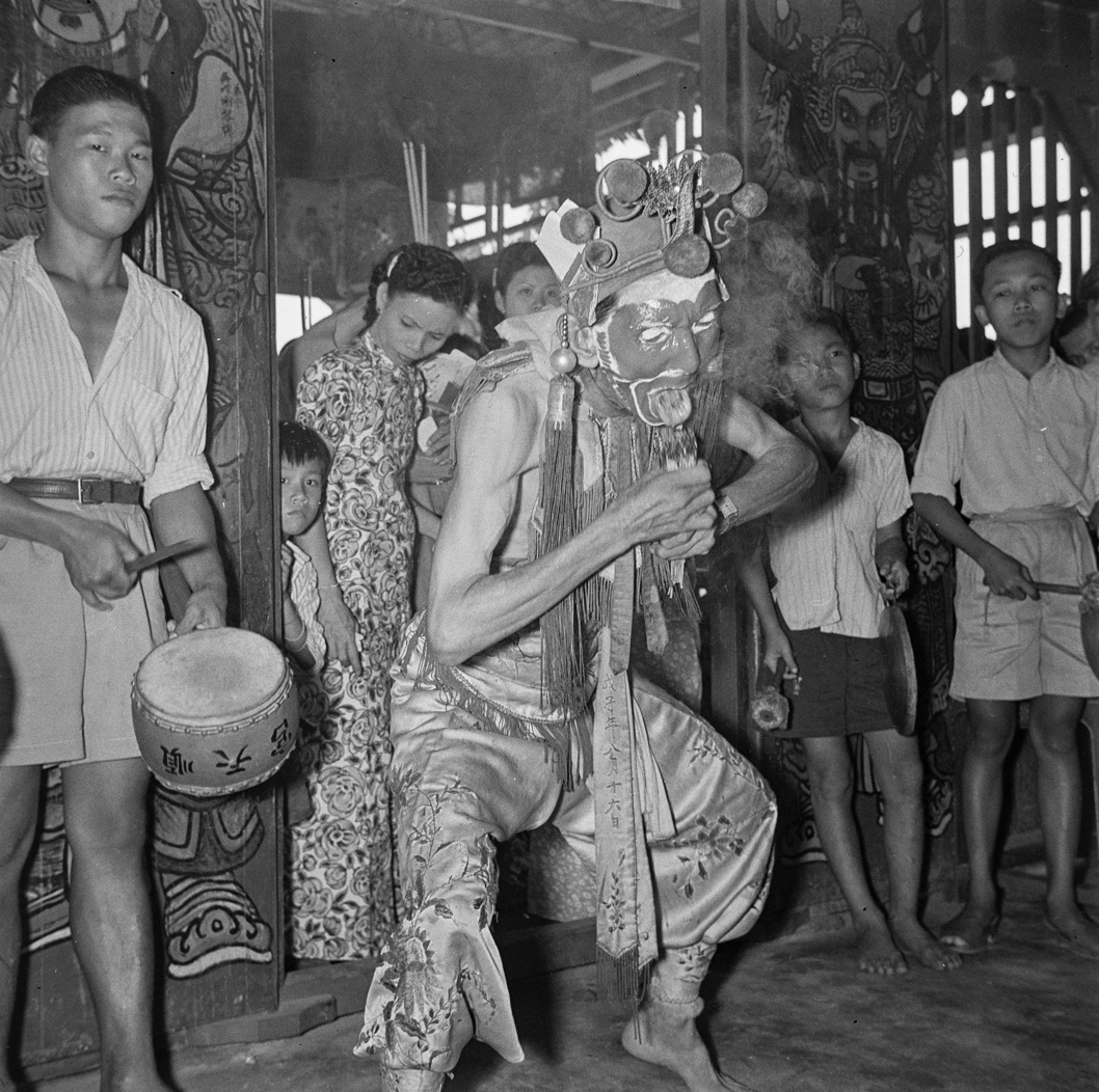 The spirit medium, an older man, has his face painted and wears a headdress and apron with silk trousers. He is holding a bundle of burning incense sticks to his tongue while standing with knees bent in front of a doorway with participants and spectators gathered behind. A man playing a drum is to the left.