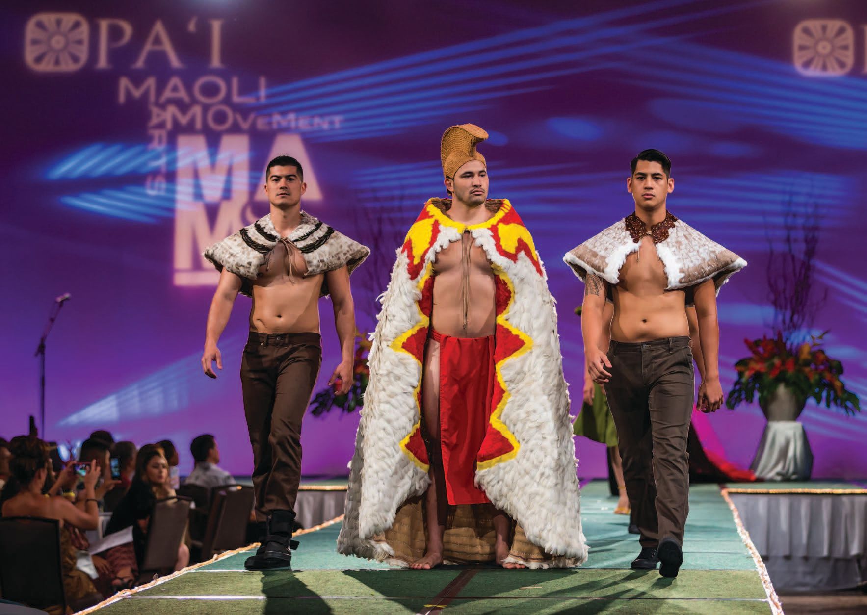 Three men walking down a catwalk at a fashion show; the background is purple and 'Pa'i Maoli Movement' is visible on the backboard. The man in the middle wears a long extravagant cloak, white with red and yellow detailing around the collar and edge, and the two men flanking him wear short shoulder capes (natural in colour) and brown trousers.