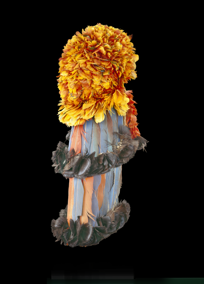 A round cap/headdress of woven fibres with red and yellow macaw feathers on the exterior with a deep fringe of longer feathers towards the back. Two rows of long blue and red feathers hanging from the back of the cap, all with plumes of dark blue/black feathers attached to the ends.