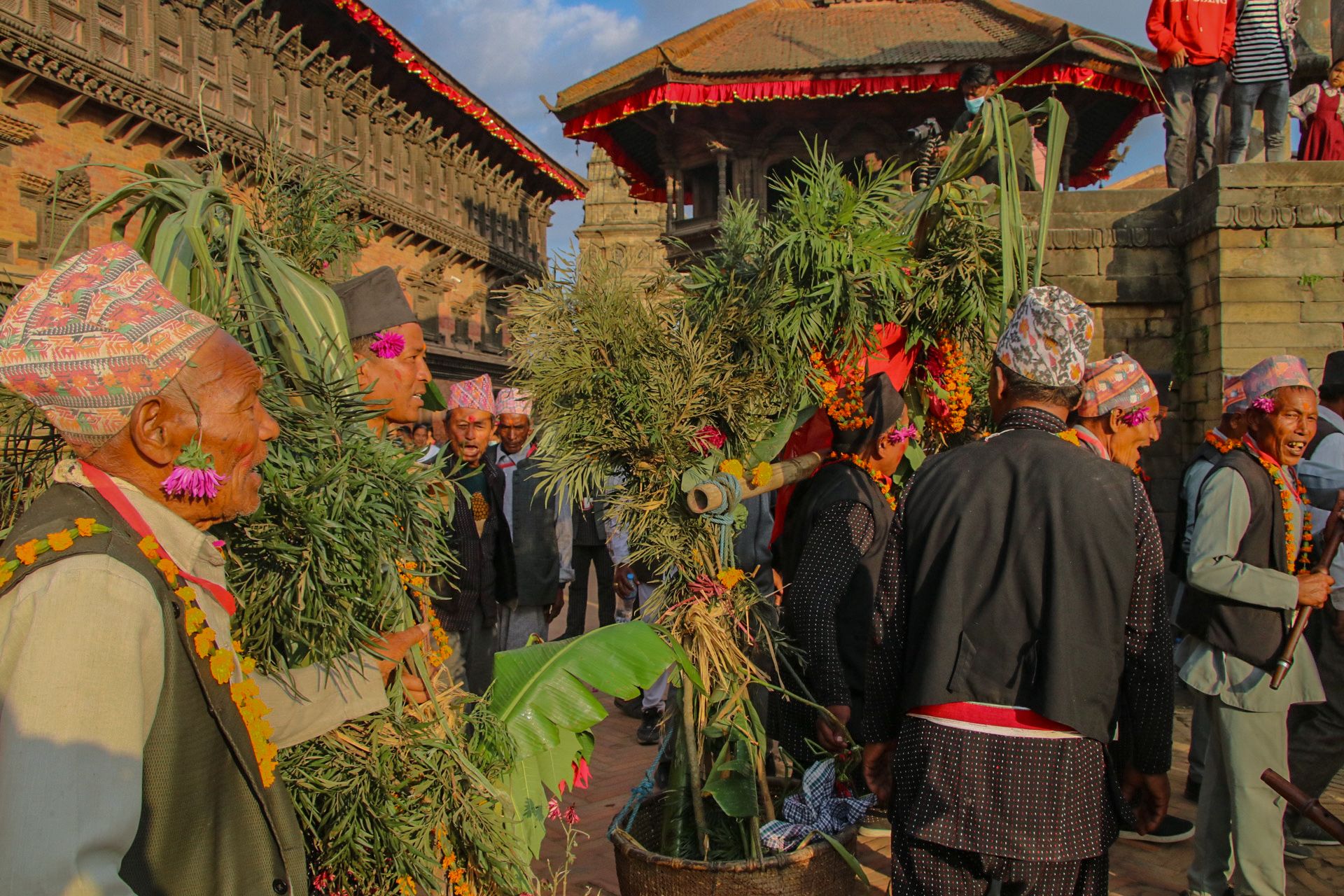 Men dressed in black and grey traditional Newari outfits with colourful caps gather in ceremony at the Bhaktapur Durbar Square carrying a variety of crop that they have recently harvested. In the backdrop we see the roof of 55 window palace with its ornate windows and panels.