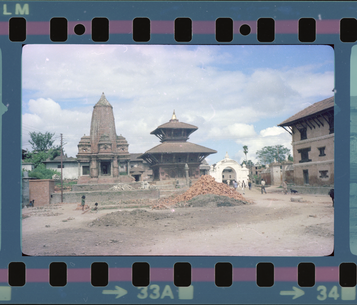 We see Construction material piled up infant of two temple, both with distinct roofs- one has a pagoda style and another has a shikhara two-tiered style roof. Both are built in wood and brick, and the constructional material too consist of brick and what seems like sand. In the distance we also see the arched white gateway of the Durbar Square. 