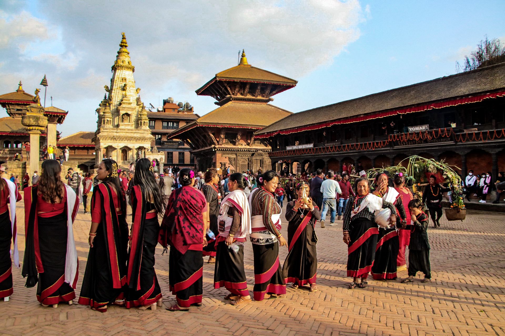 Women dressed in a traditional Newari black saree with a red border are gathered in the Durbar Square. Some men are seen carrying harvested crop, while the women carry golden pots of water. In the background are temples built in brick and wood, and a single temple built in white stone. Each temple has a distinct and ornate roof that is clearly visible in the image. 
