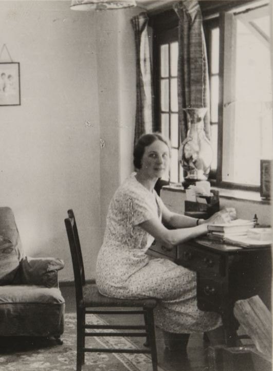 A portrait of Margaret Williamson wearing a printed dress sitting at her desk inside her office at the British Residency in Gangtok, where she and her husband, Frederick Williamson, lived during his posting as Political Officer. The room contains a dresser, armchair, desk chair, desk and rugs on the floor. There are framed pictures hanging on the wall and standing on the dresser in front of a jade screen.