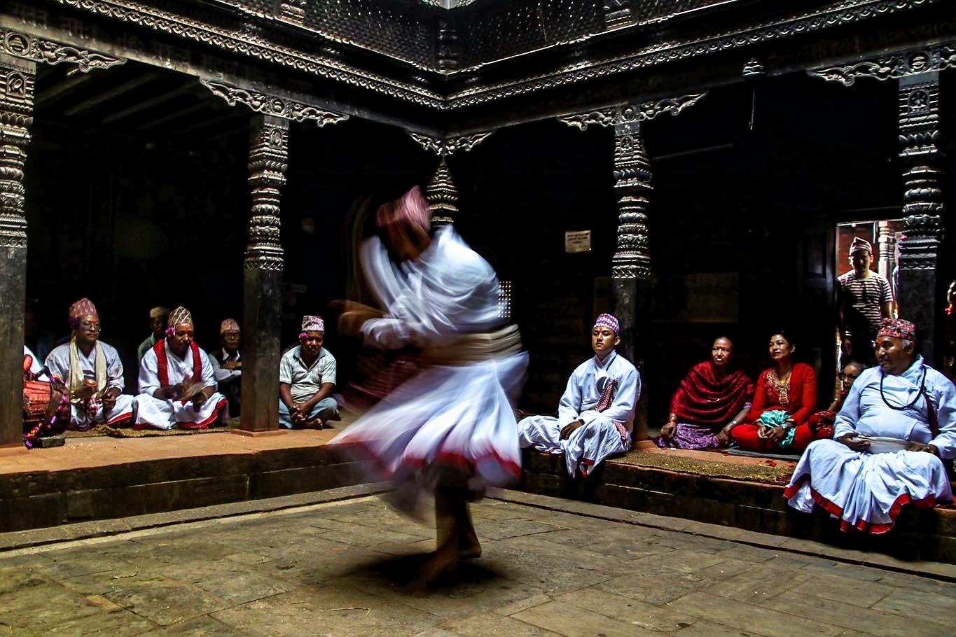 A dancer dressed in a traditional white skirted gown is captured moving passionately in a dark inner sanctum of a temple with exquisite terracotta pillars. Am audience comprised of dancers dressed similarly, musicians and women look on.