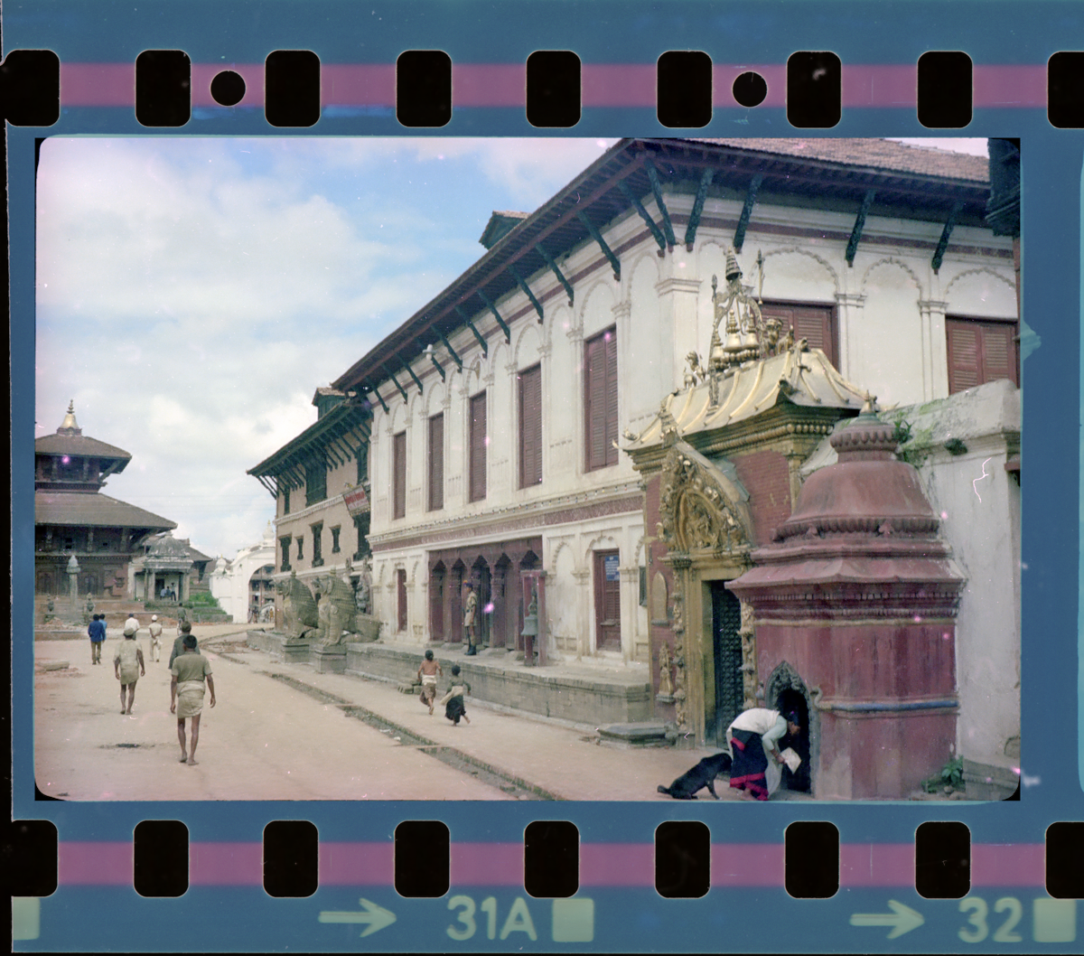 A photograph of the important structures in the Durbar Square - to the right is a gateway to a temple with a torn gilded in gold and copper with intricate metalwork, beside which is a the Palace Quarters built in white stone with wooden windows and doors, and in the distance is the National Art Museum built in brick, and temple built in brick and wood with a two tiered roof. This is a typical image of the Bhaktapur Durbar Square and we see also see a woman worshipping at a shrine, people walking, children praying, and a soldier guarding the palace quarters' gates.