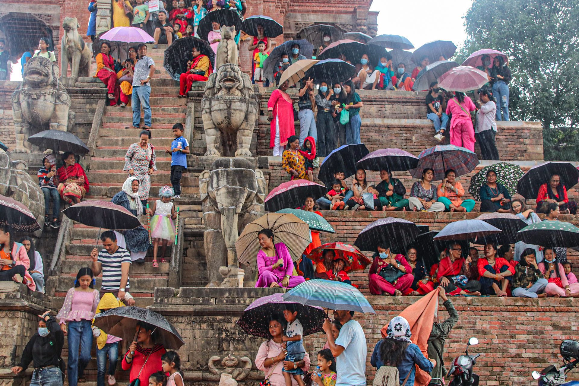 People dressed in colourful clothes with umbrellas gather on the steps of a temple to take shelter during the rain. They sit around stone sculptures of elephants and other mythical creatures bordering the main stairway that leads up to the temple. 