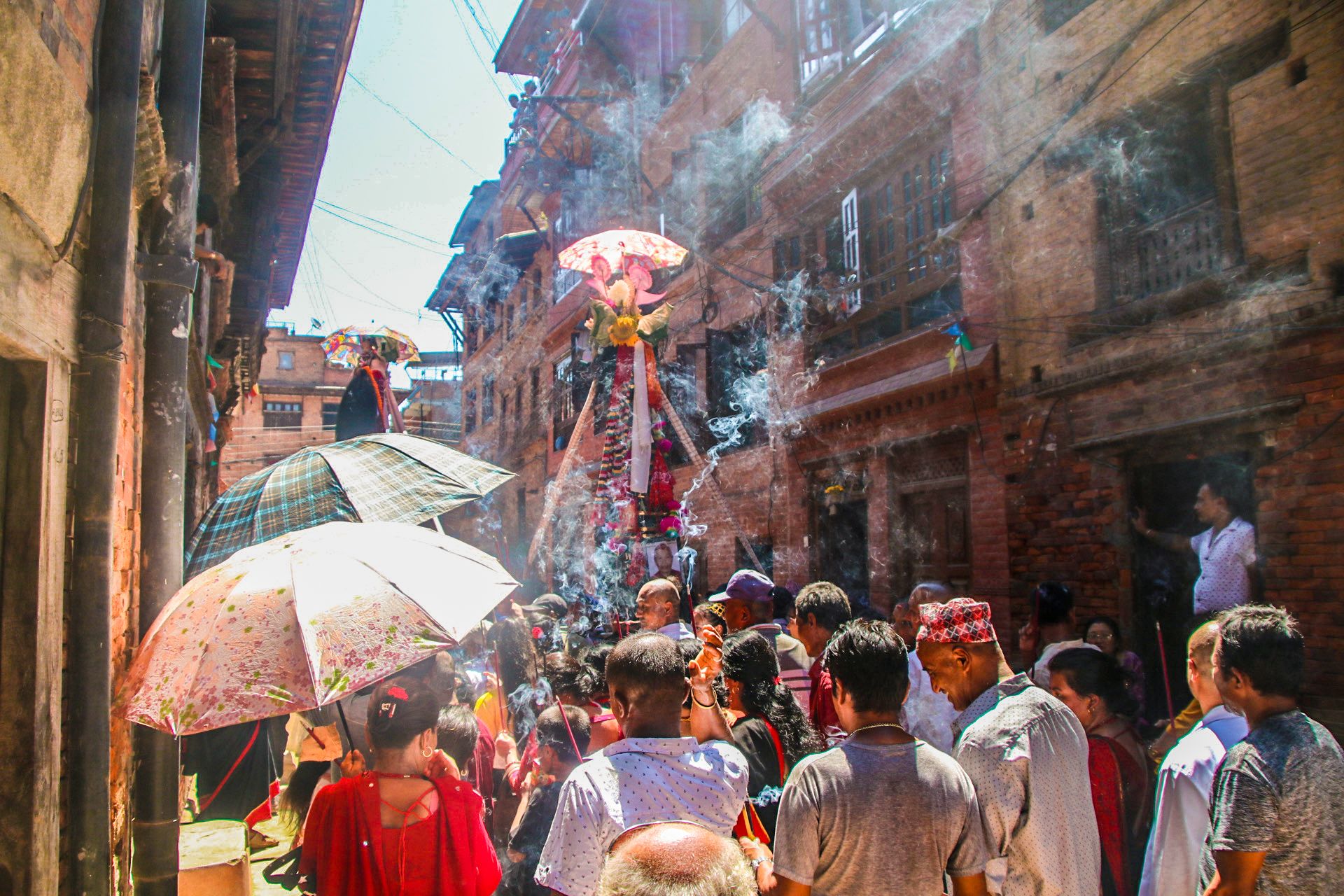 People are seen in procession carrying an effigy of the cow that represents a deceased family member. The effigy is shaped conically and is made of wooden poles and an umbrella on top, and has flower garlands surrounding a photograph of the deceased that is mounted on the intersection of the poles. People are burning incense in honour of the effigy, and some are carrying umbrellas since it's sunny. They walk through a lane in a neighbourhood surrounded by houses built in brick and wood on either side.