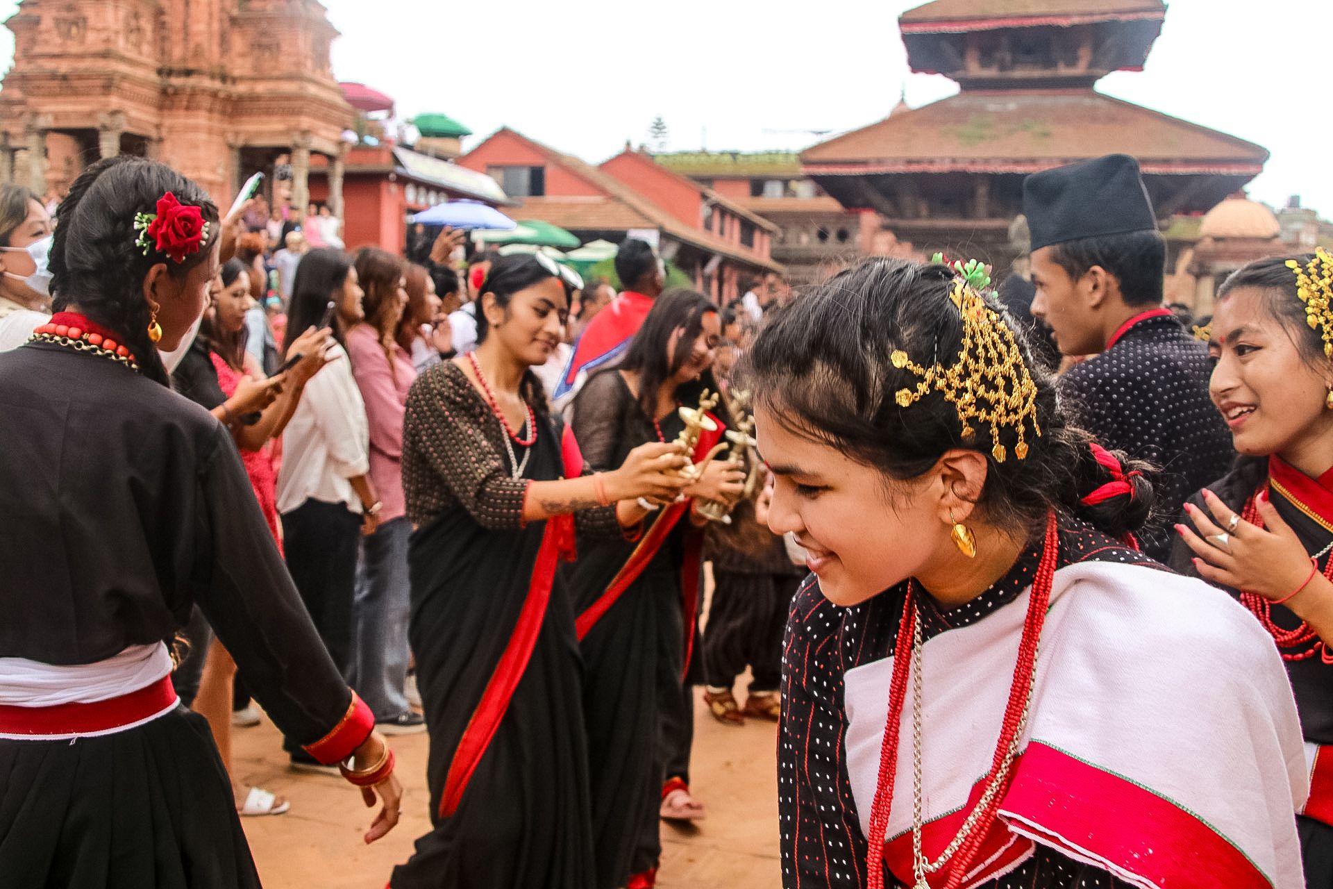Young residents, mostly women, are seen dancing in a traditional Newari black saree with a red border, decked in gold jewellery. Some women have ritual instruments made in gold in their hands.In the background we see the roof of a temple in brick, and a few more brick buildings in the distance.