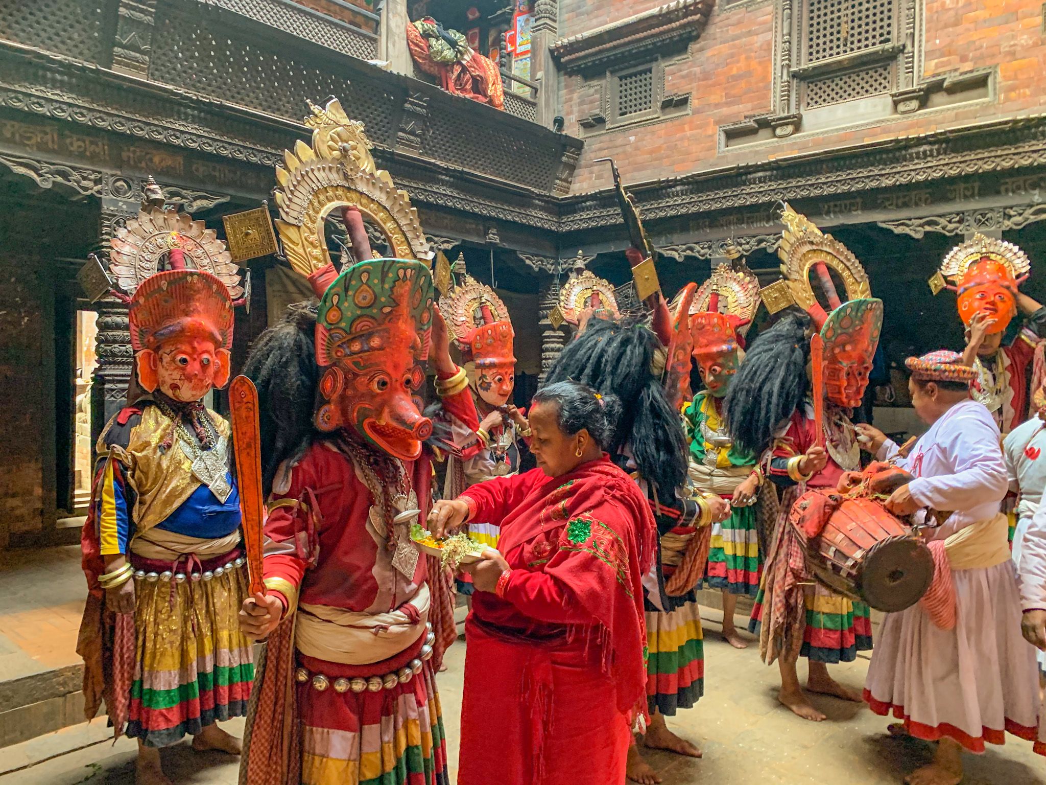 Men(NavaDurga dancers) wear elaborate fierce masks representing demonic goddesses. Here the masks are being blessed by a woman who is a senior member of the group. They are all gathered at the temple of the NavaDurgas and in the photograph we see a glimpse of the beautiful wooden panels and ornate pillars of the temple. 