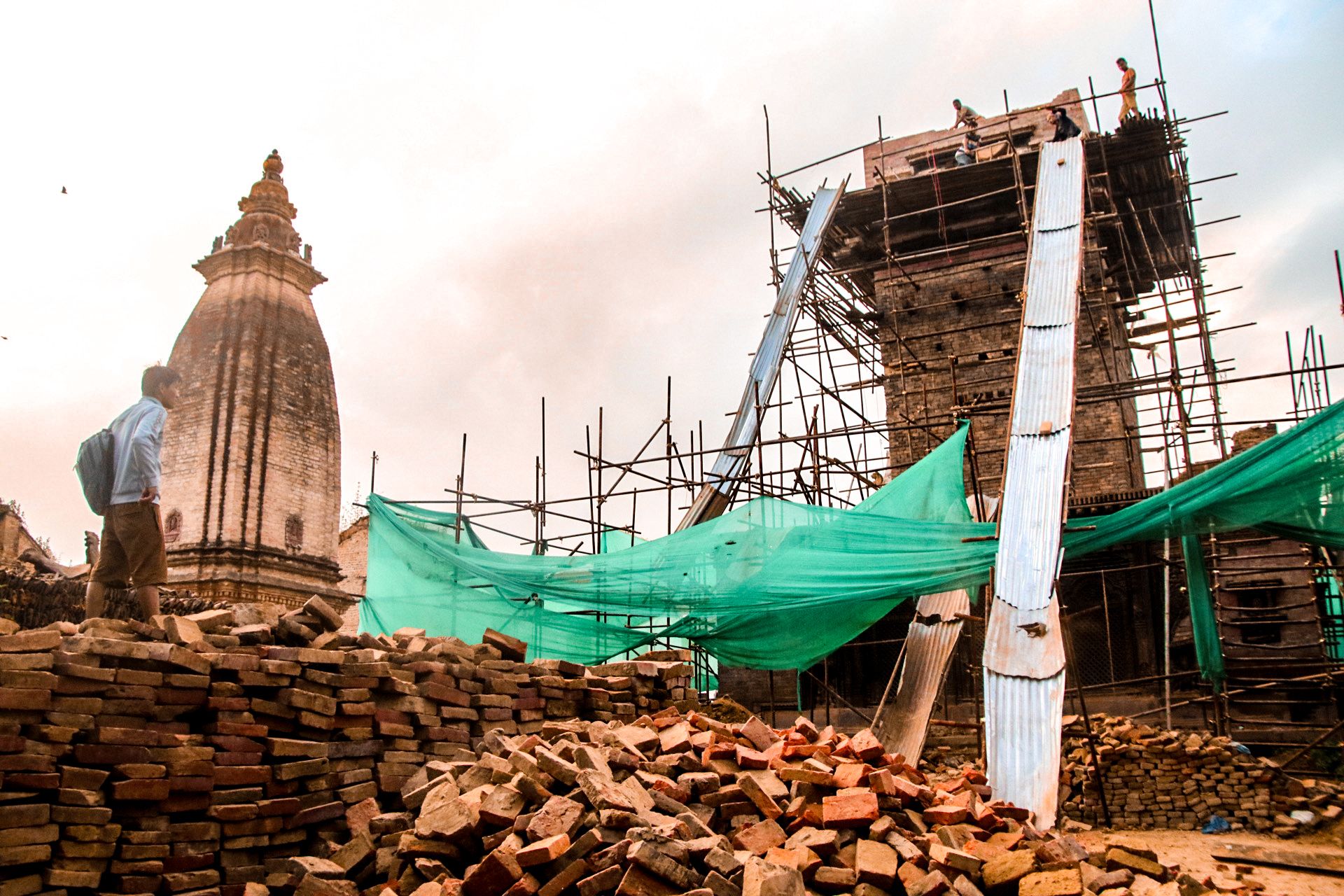 We see construction material including brick and wooden panels piled up infront of a temple that is under construction. There are people working on the roof of the temple that is surrounded in bamboo scaffolding. 