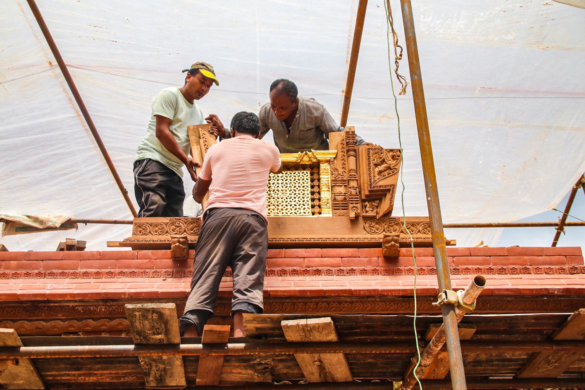 People fix a gold-gilded window set in a wooden frame carved intricately with HIndu mythological and religious motifs. The window is being fixed on to the first floor of a temple that is under construction and we see bamboo scaffolding around the temple. 