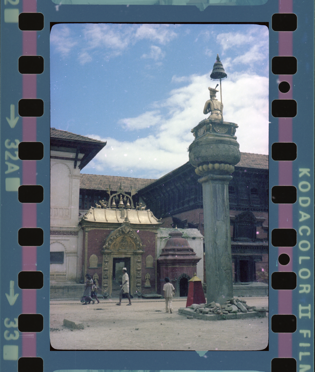 Here we see a gateway that leads to a temple and palace that aren't fully visible in the photograph. The entryway has a golden arch and roof, with inscriptions in Newari, iconographic motifs and scriptures all carved in metal. In front of the temple gateway is a pillar with a mythological figure sitting atop, and beside the temple is small shrine.