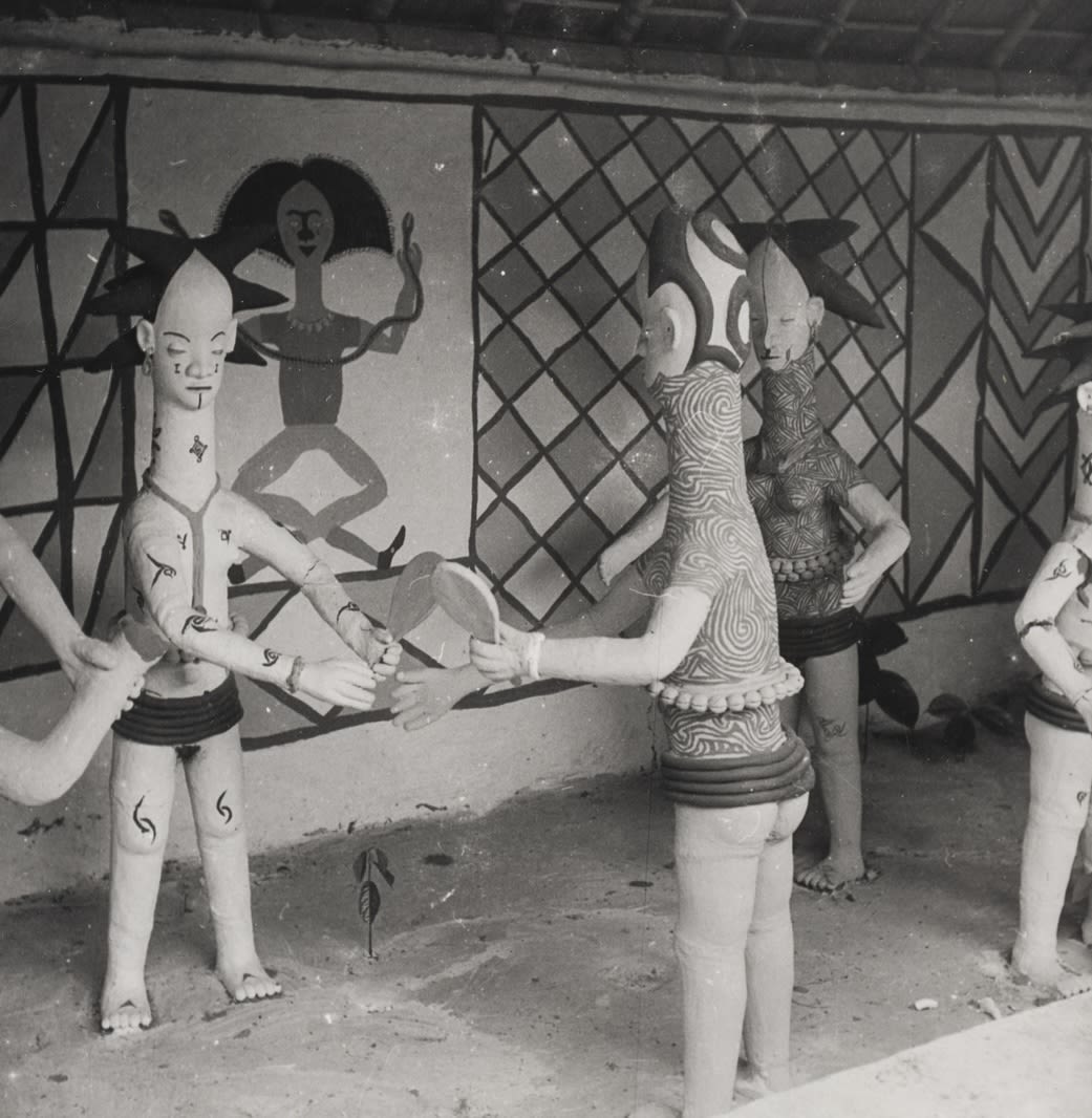 Group of clay figures arranged in front of patterned wall paintings, and holding mirrors and adornments in their hands.