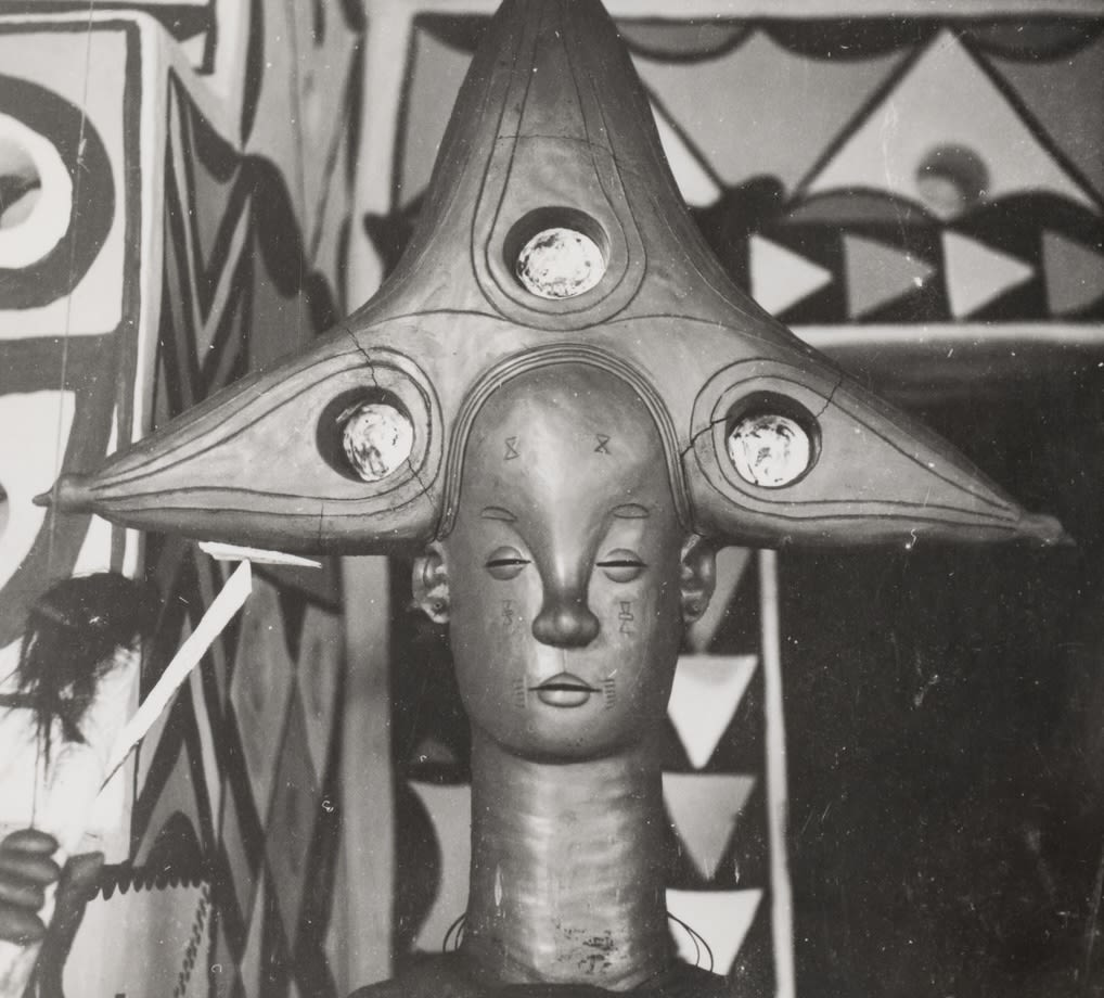 Close-up of a sculpted head with an elaborate, triangular headdress featuring circular insets, photographed against a painted geometric backdrop.