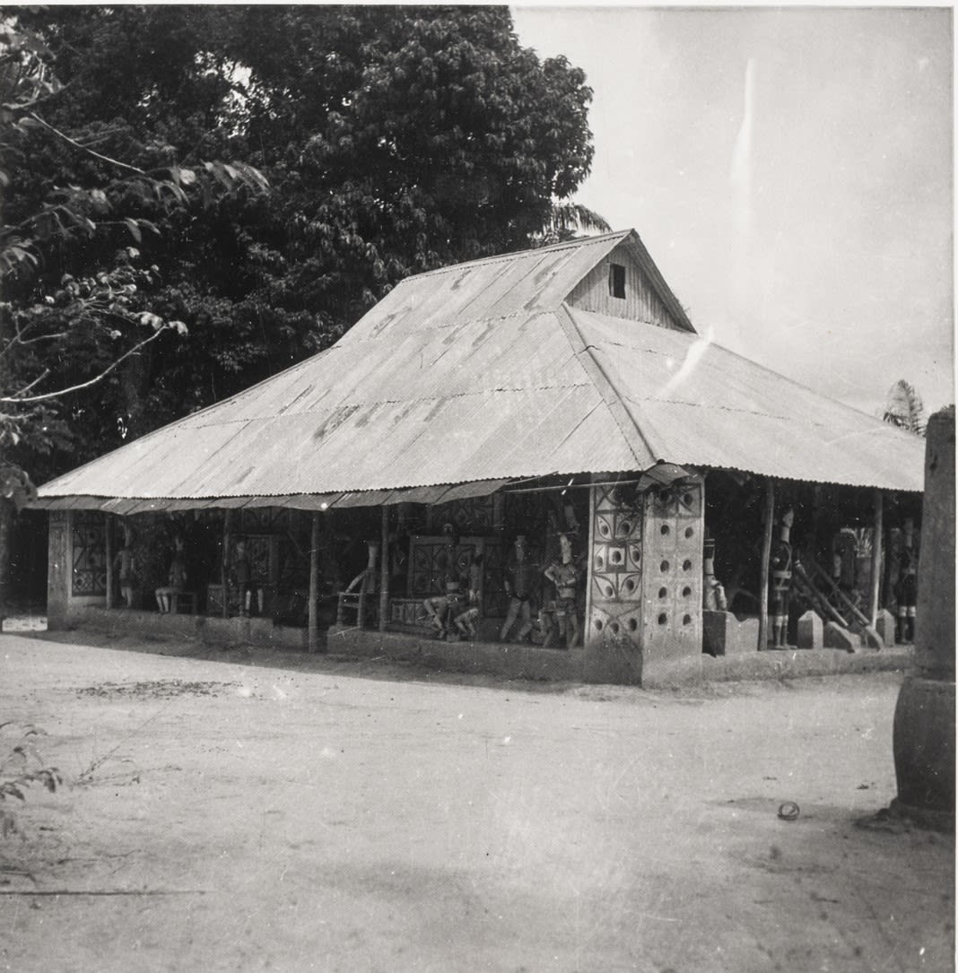 Black-and-white photograph of a long, low building with a corrugated roof and painted pillars, surrounded by trees.