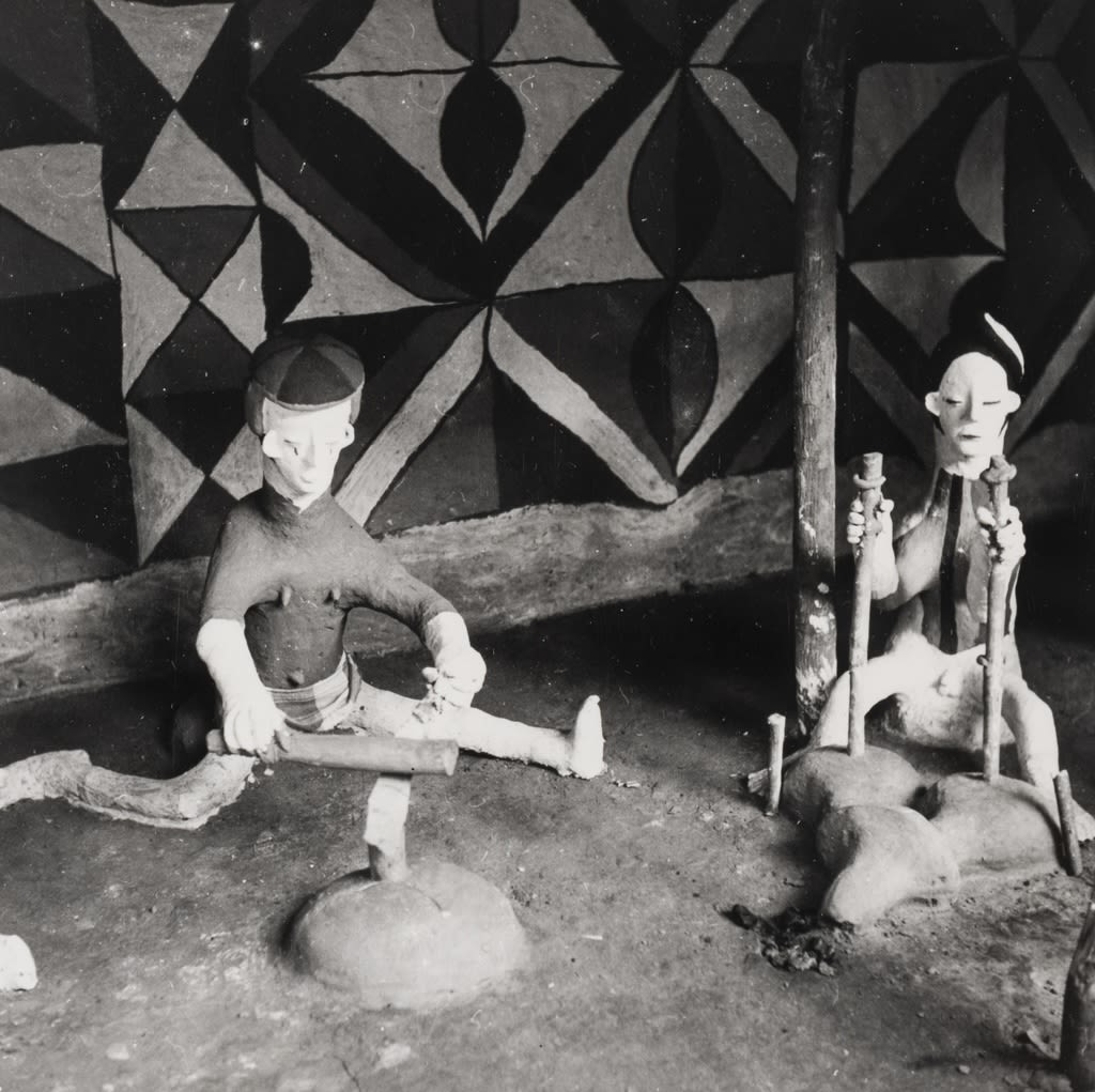 Two clay figures posed indoors against a wall painted with bold geometric patterns; one figure sits on the ground using a tool, while another kneels holding upright objects.