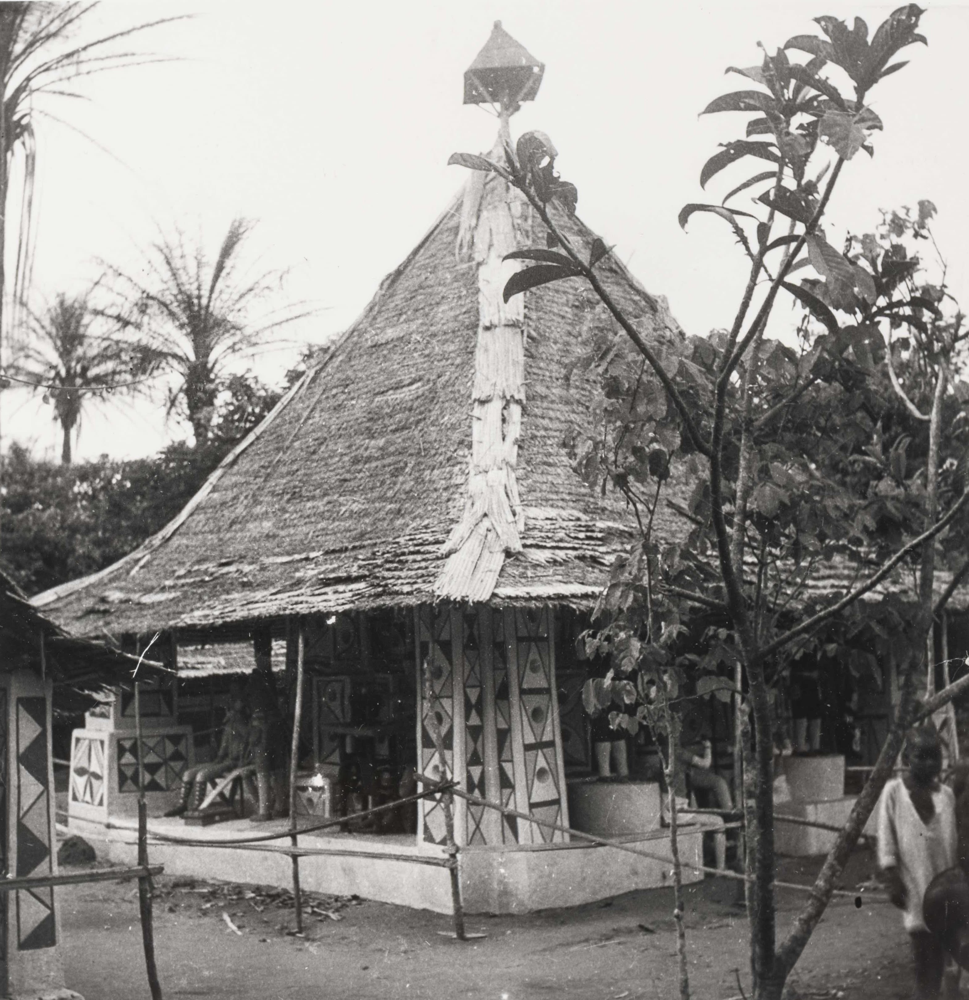 Black-and-white photograph of a thatched-roof building with painted geometric designs on the walls and pillars; trees and a person visible at the edge.