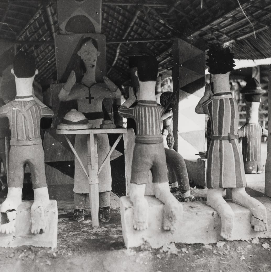 Clay figures arranged around a central female figure dressed like a nun and stood behind a small table; the surrounding figures kneel on a step facing the female figure.