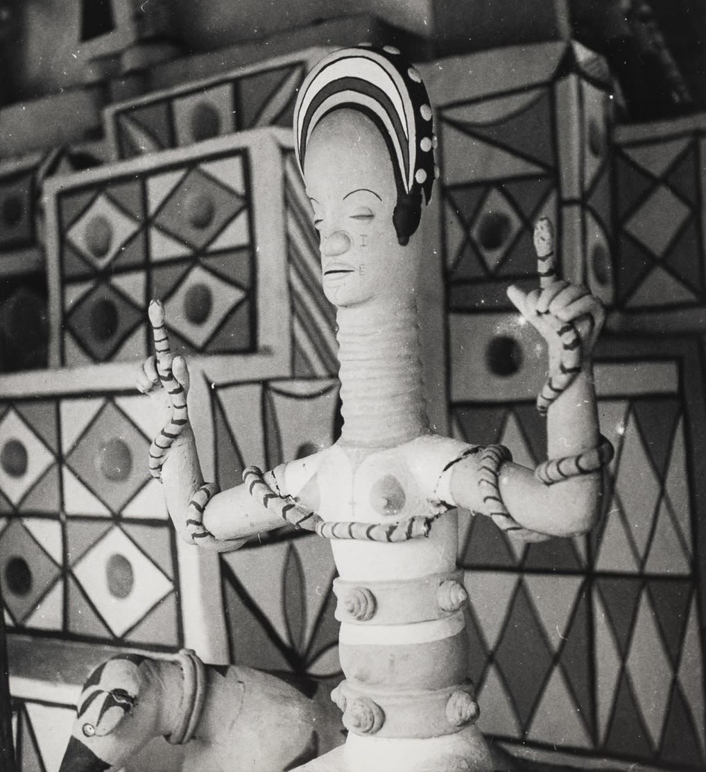 Clay female figure with elongated neck and raised arms, with snakes coiled around her arms and torso, set against a painted geometric background.