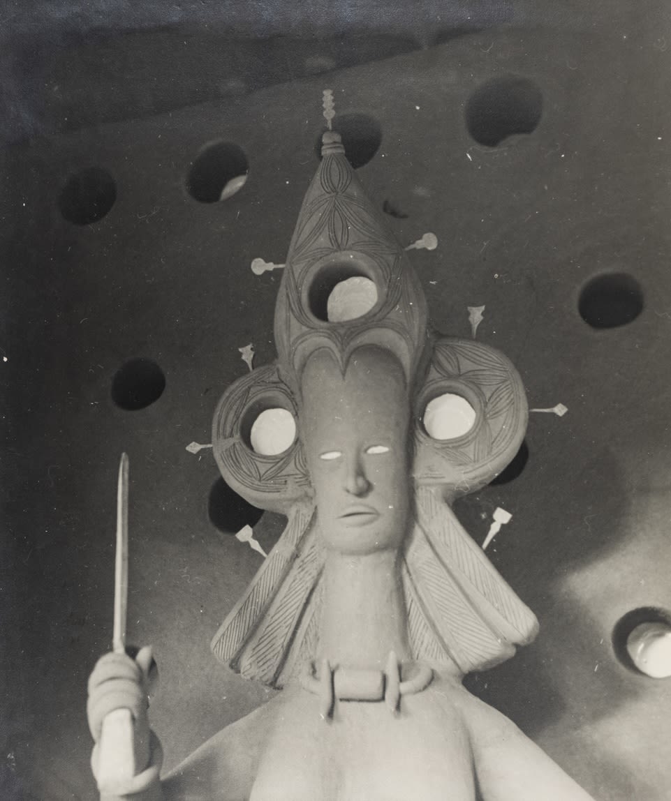 Black-and-white photograph of a clay figure inside an Igbo mbari house, with an elongated face, elaborate headdress, circular wall recesses behind, and one arm raised holding a knife.