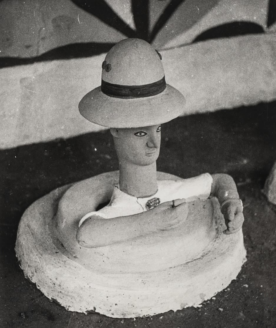 Clay figure wearing a brimmed hat, seated inside a circular basin-like form, with arms bent forward.
