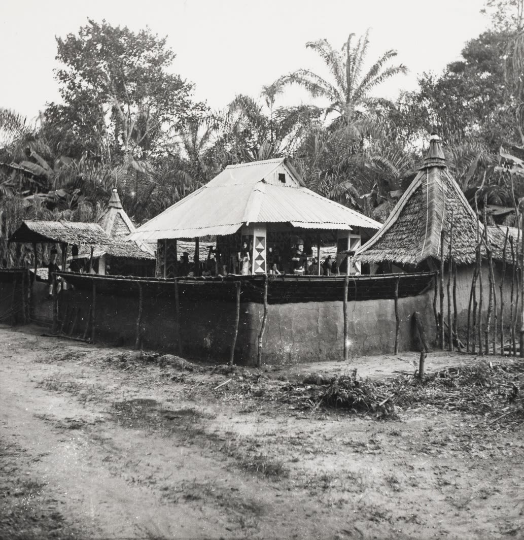 Exterior view of a circular or semi-circular structure enclosed by a low wall, with a corrugated metal roof and painted panels, surrounded by palm trees and dense vegetation.