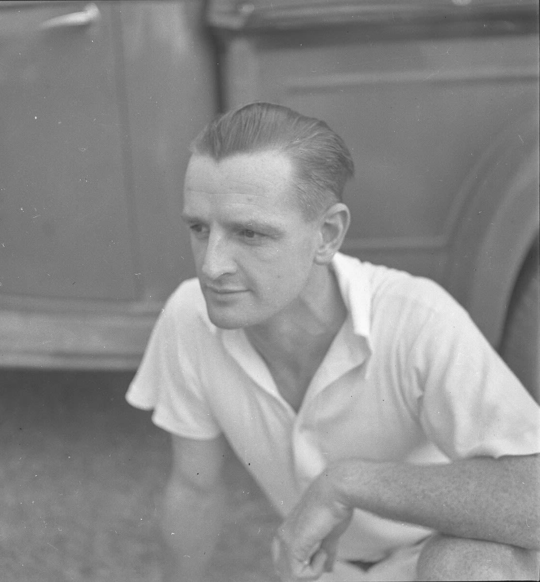Black-and-white portrait of a man crouching beside a vehicle, wearing a light short-sleeved shirt and looking to the left.