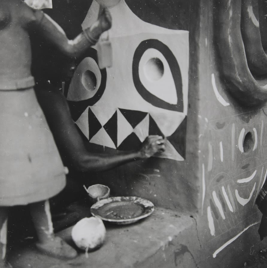 Person painting a bold, geometric face motif on the exterior wall of a building; bowls of pigment rest on the ground beside the wall.