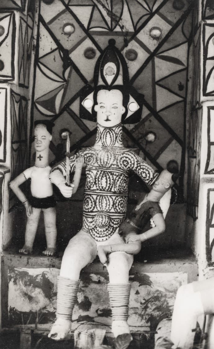Seated clay figure holding a child on one knee, framed by painted geometric panels; a smaller standing figure appears behind to the left.