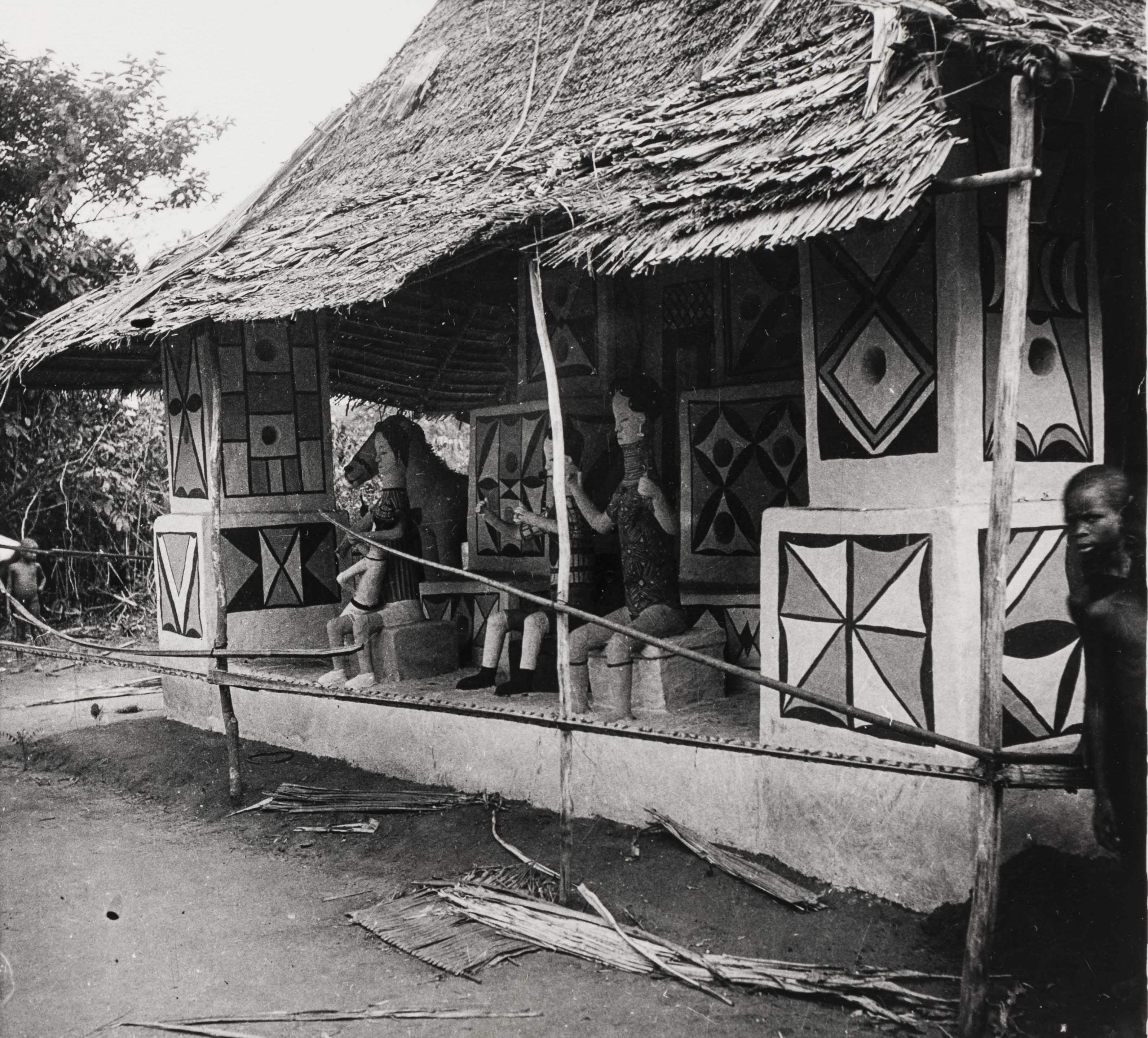 Exterior view of a painted shrine with a thatched roof; sculpted figures sit along the front platform while a person stands at the right edge of the frame.