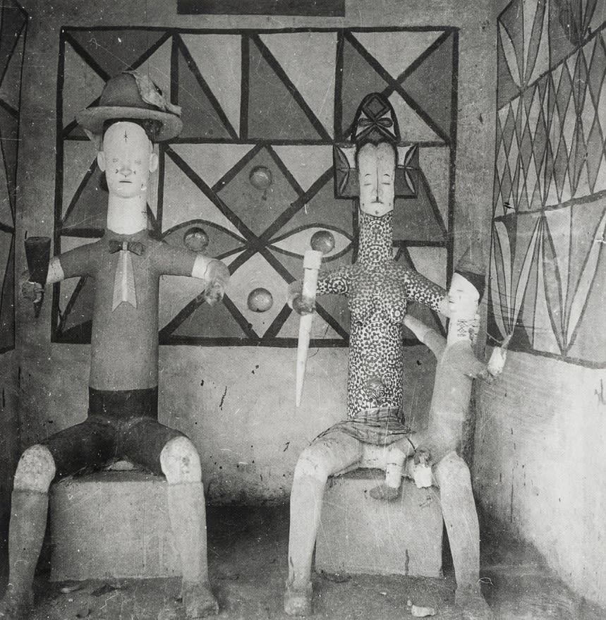Black-and-white photograph of two large seated sculpted figures inside a small painted room, with geometric wall patterns behind them.