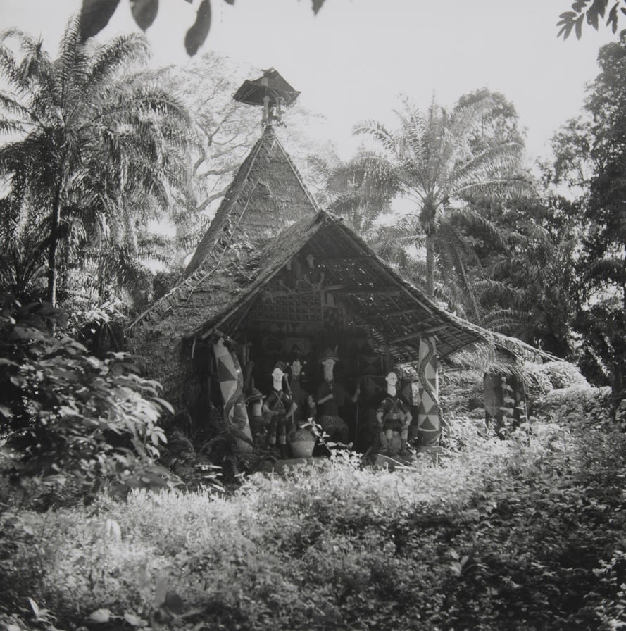 A thatched structure set within forest vegetation, with carved or sculpted figures arranged inside the open front beneath a steeply pitched roof.