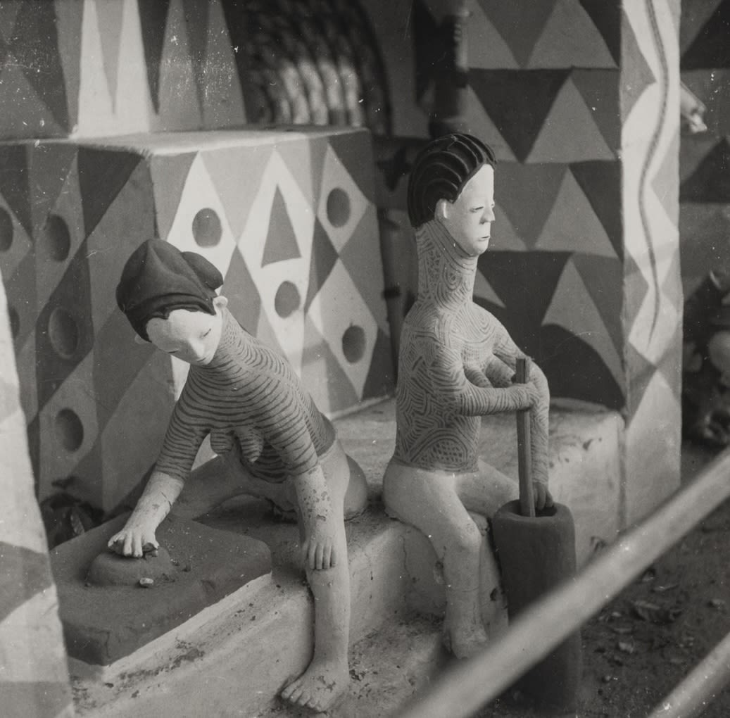 Two clay figures sit side by side on a raised platform inside a painted structure; one leans forward with hands on the ground, while the other sits upright holding a long pestle set into a mortar.